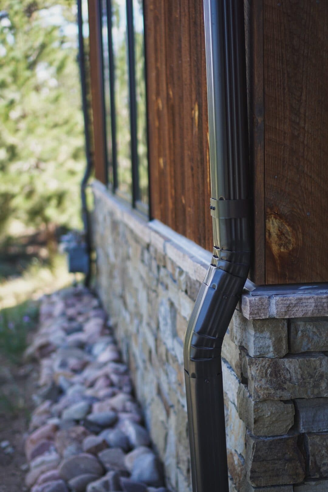 Bronze aluminum downspout discharging onto a river-rock drainage bed away from the foundation of a Colorado mountain home, installed by Green Brothers Gutters.